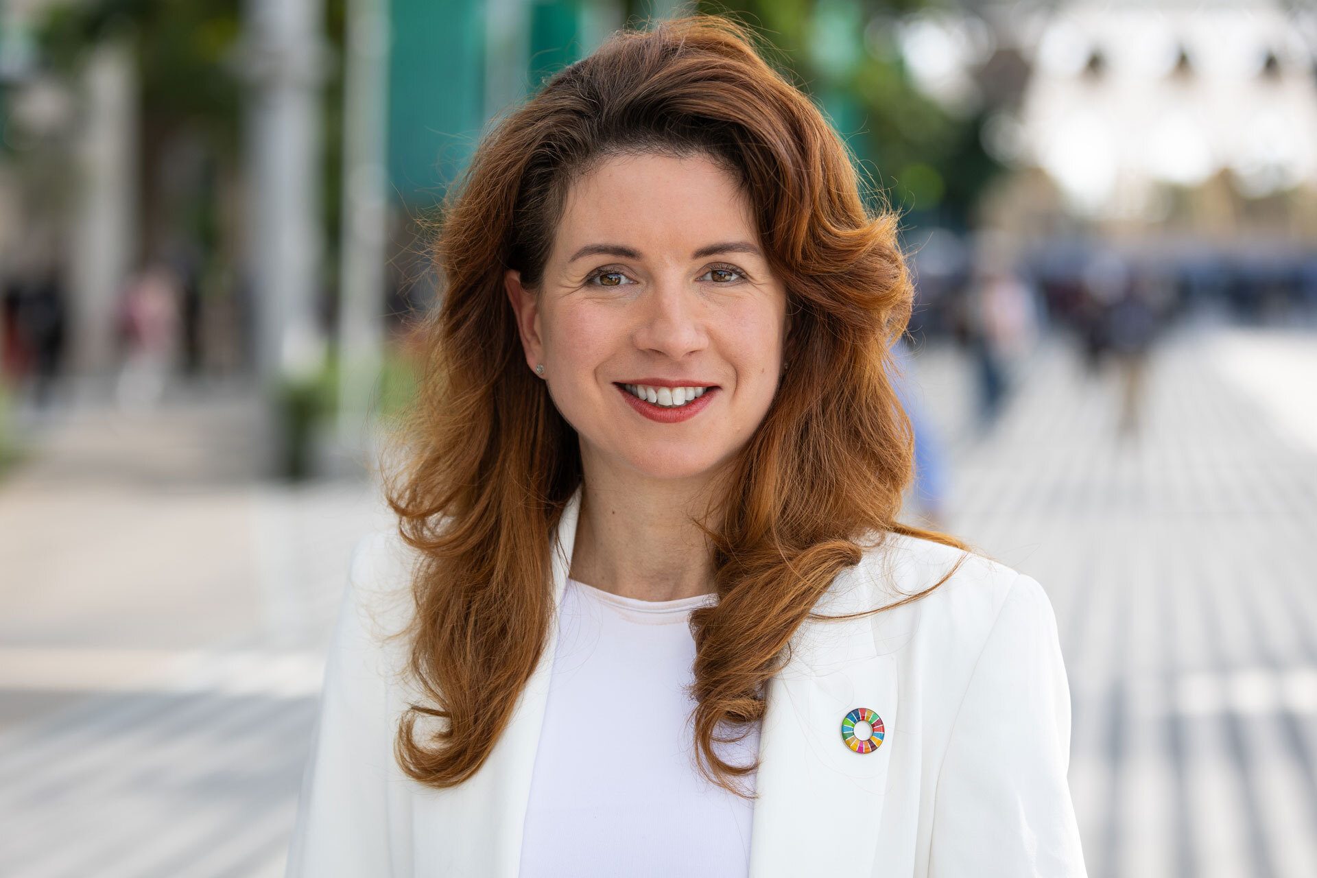 Red-haired woman in white blazer with SDG pin, smiling outdoors.