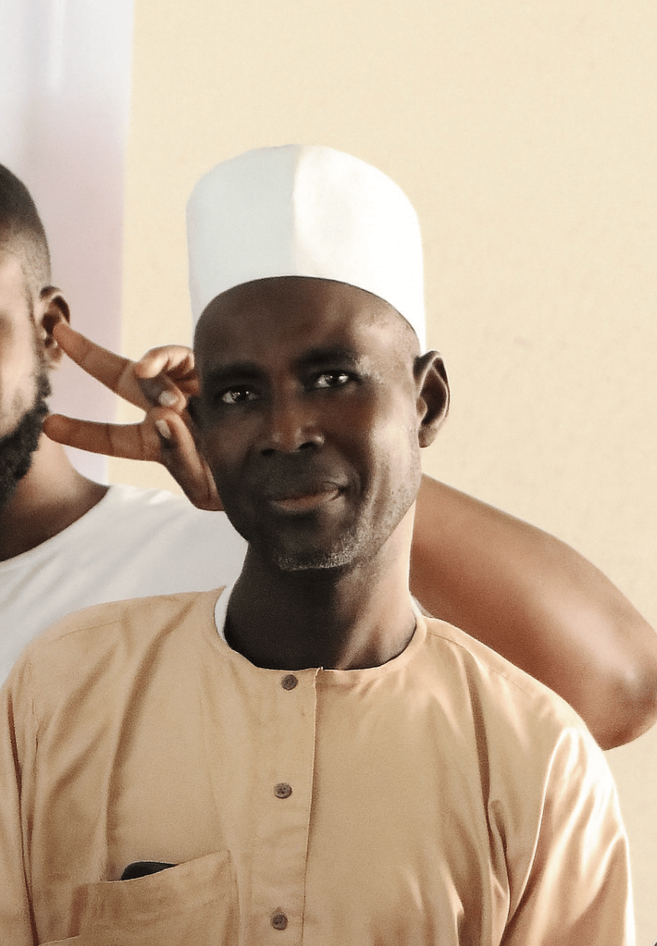 Smiling man in a white cap and peach shirt, with a hand making a peace sign near his ear.