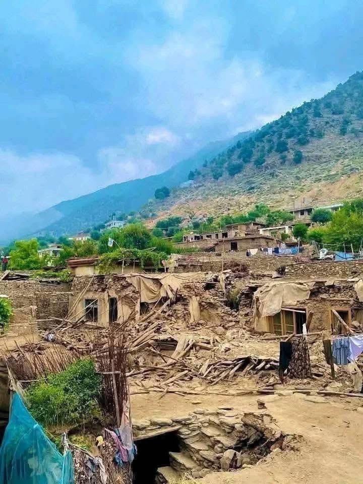 Devastated mountain village with crumbled homes and debris against a backdrop of green mountains under a cloudy sky.