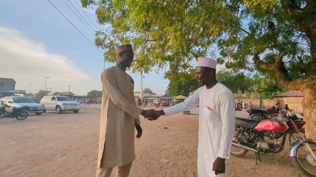 Two men in traditional attire shake hands outdoors under a tree, symbolizing greetings or agreement.