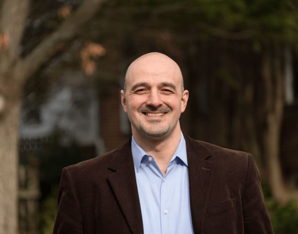 Bald man with a big smile, wearing a blue shirt and brown jacket, standing outdoors.