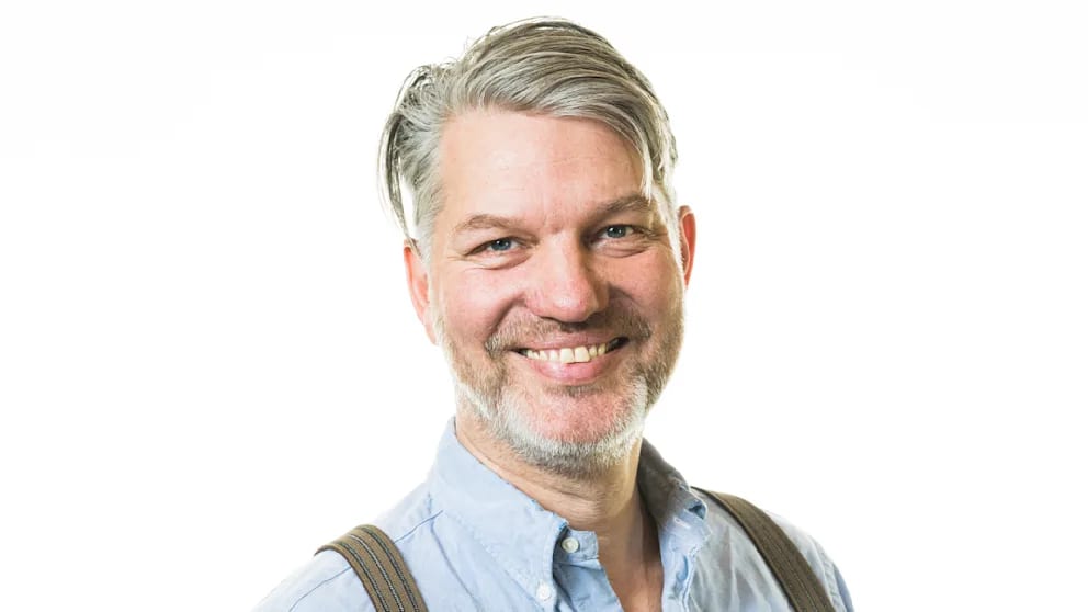 Smiling man with grey hair and beard, wearing a blue shirt and suspenders, on a white background.