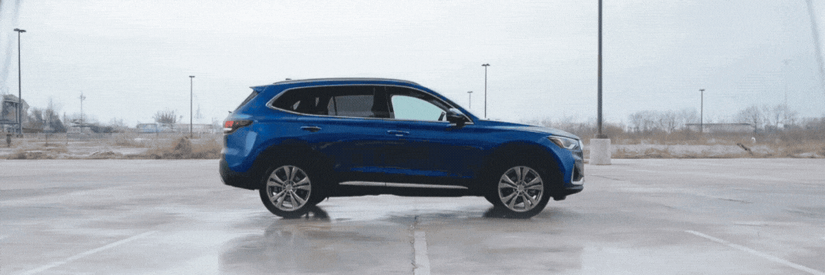 A blue SUV drives across a wet paved surface, splashing water from its tires under an overcast sky.