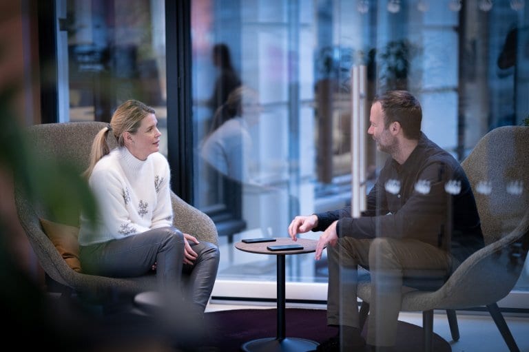 Man and woman sitting and talking in chairs by a reflective office window.