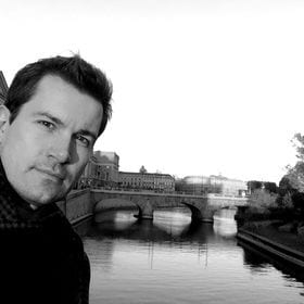 Man's face in black and white, with a canal, bridge, and city buildings in the background.
