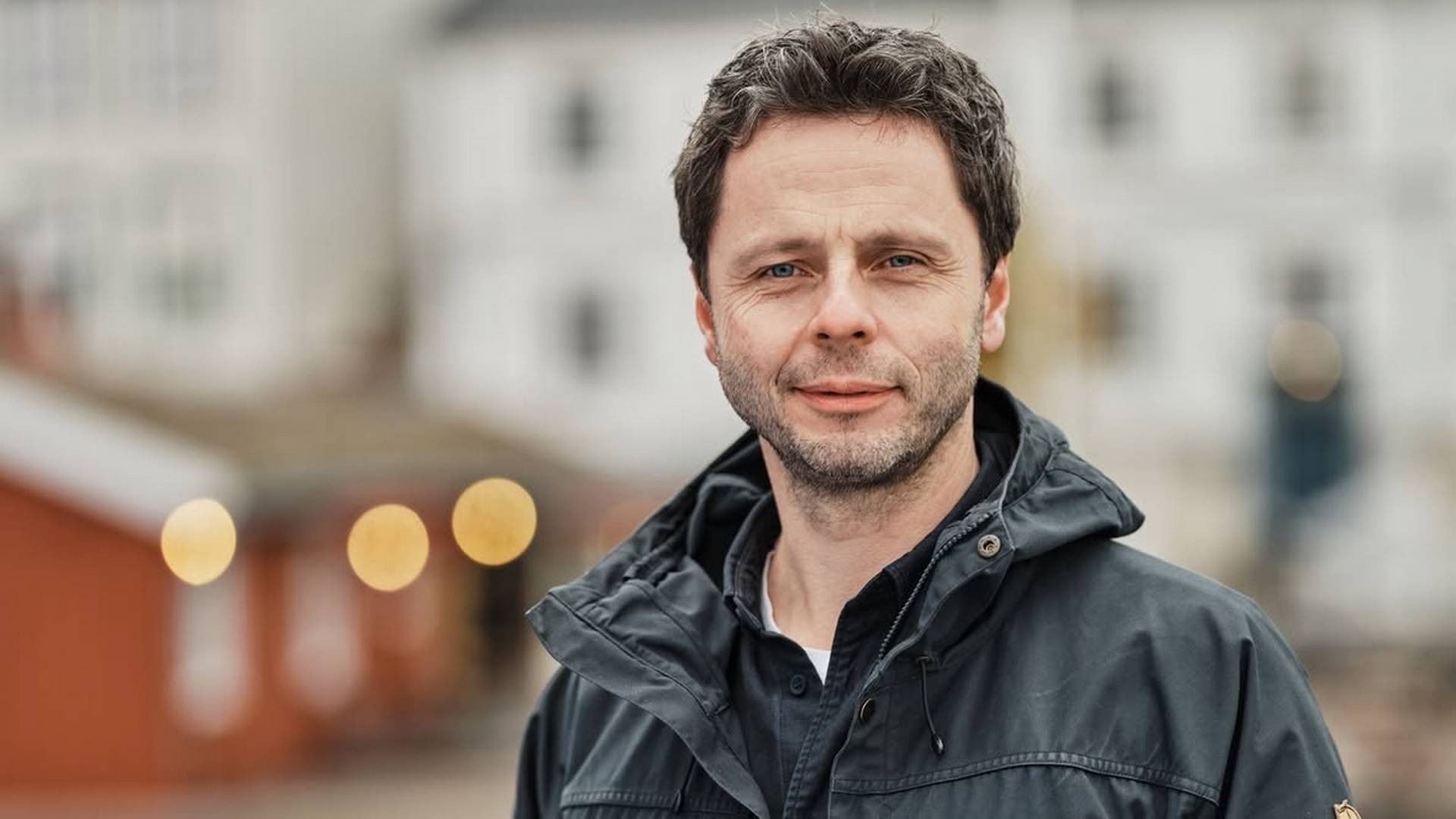 Man with dark hair and blue eyes, wearing a dark jacket, looking at the camera outdoors. Blurred background.