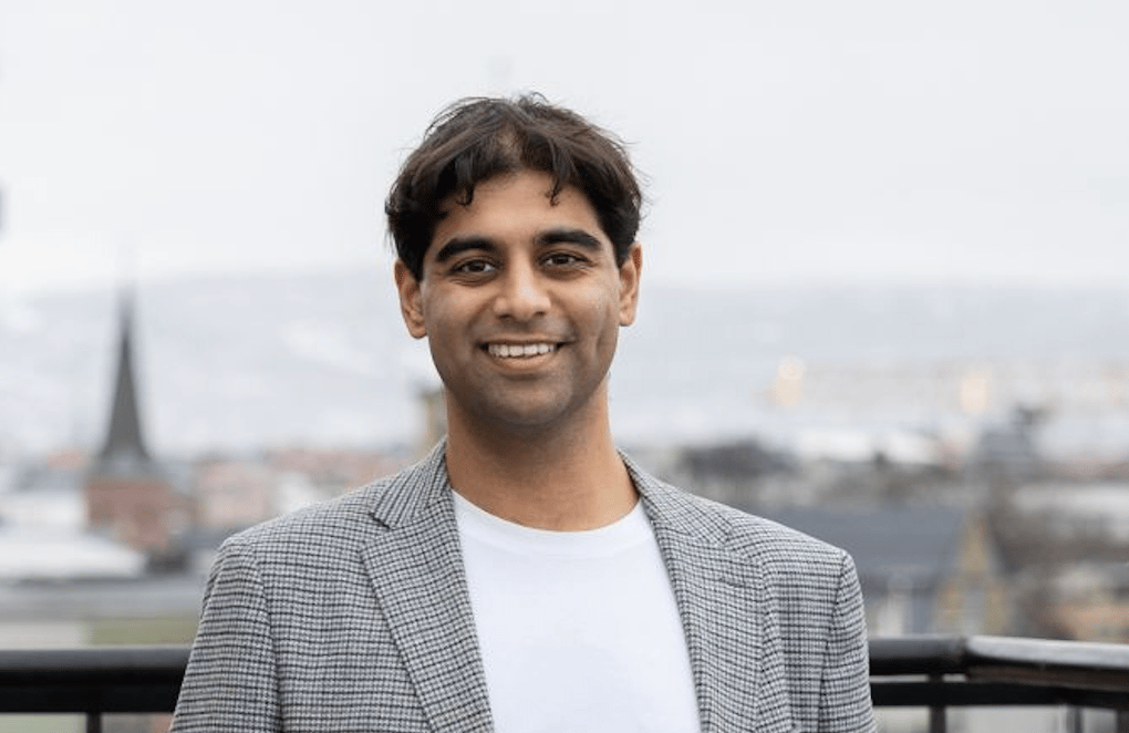 A smiling man with dark hair in a grey blazer and white t-shirt, standing outdoors with a city view.