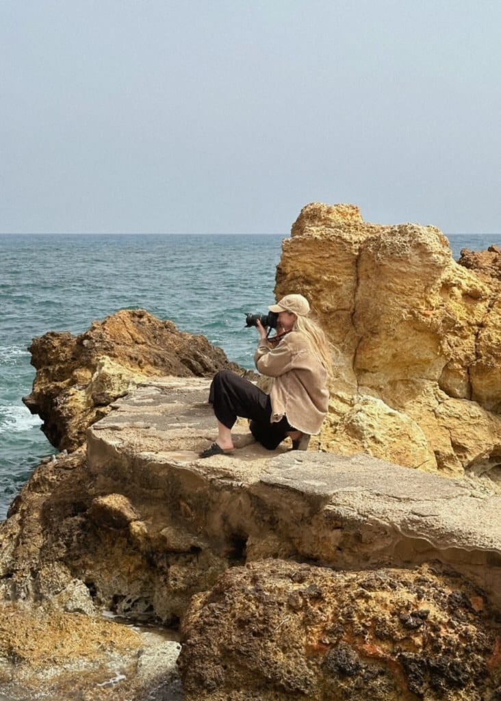 Blonde woman in cap crouched on rocks by the sea, taking a picture with a camera.