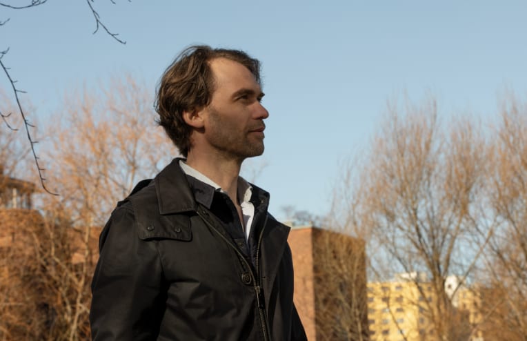 A man in a dark jacket with a beard looks contemplatively to the right against a bright blue sky.