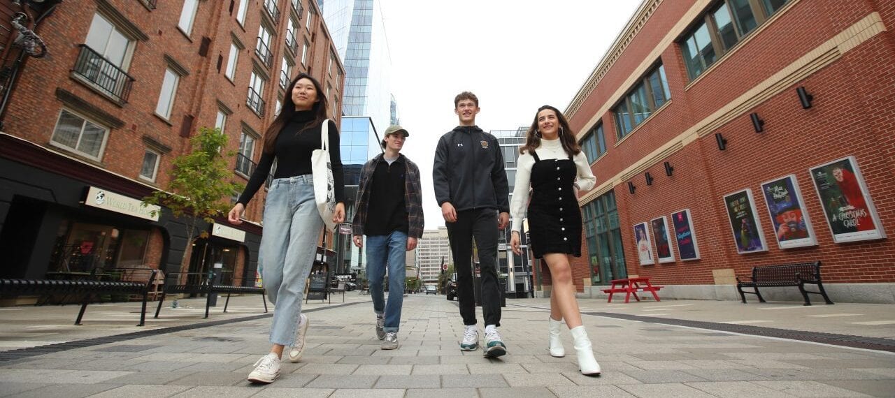 Four young people walking together down a city street lined with brick buildings.