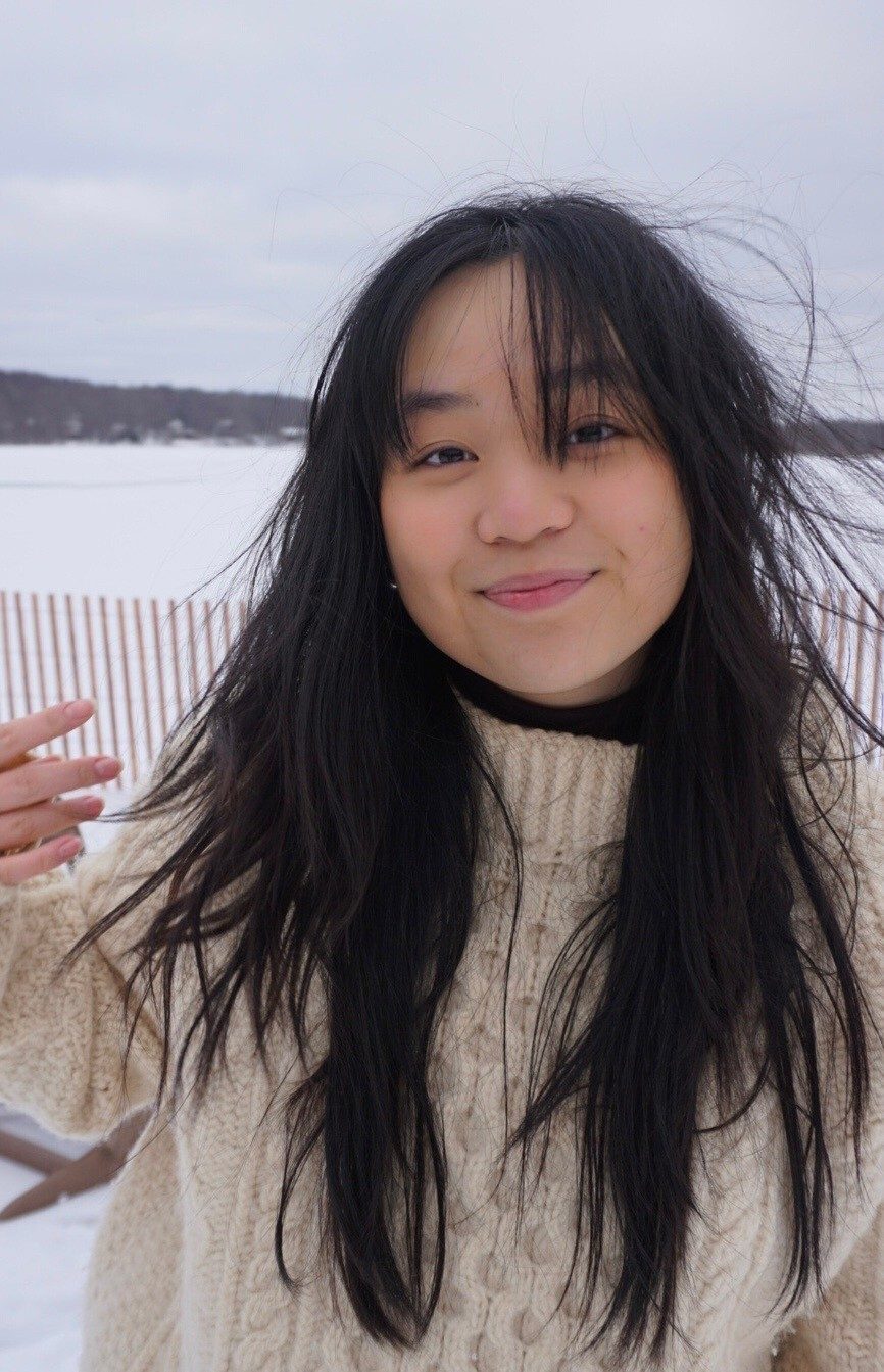 Smiling young Asian woman with windswept dark hair in a cream sweater, in a snowy winter scene.