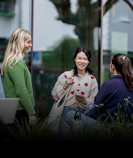 Three young women chat outdoors, one in a heart-patterned cardigan holds a phone.