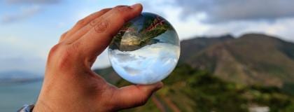 Hand holds crystal ball reflecting inverted mountains, sky, and sea.