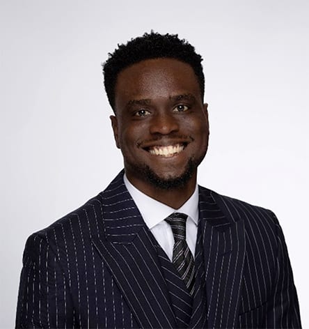 A smiling man with short dark hair in a pinstripe suit and tie looks directly at the viewer.