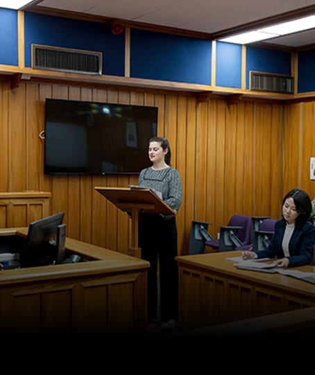 Two women in a courtroom-style room: one speaks at a podium, the other sits and writes.