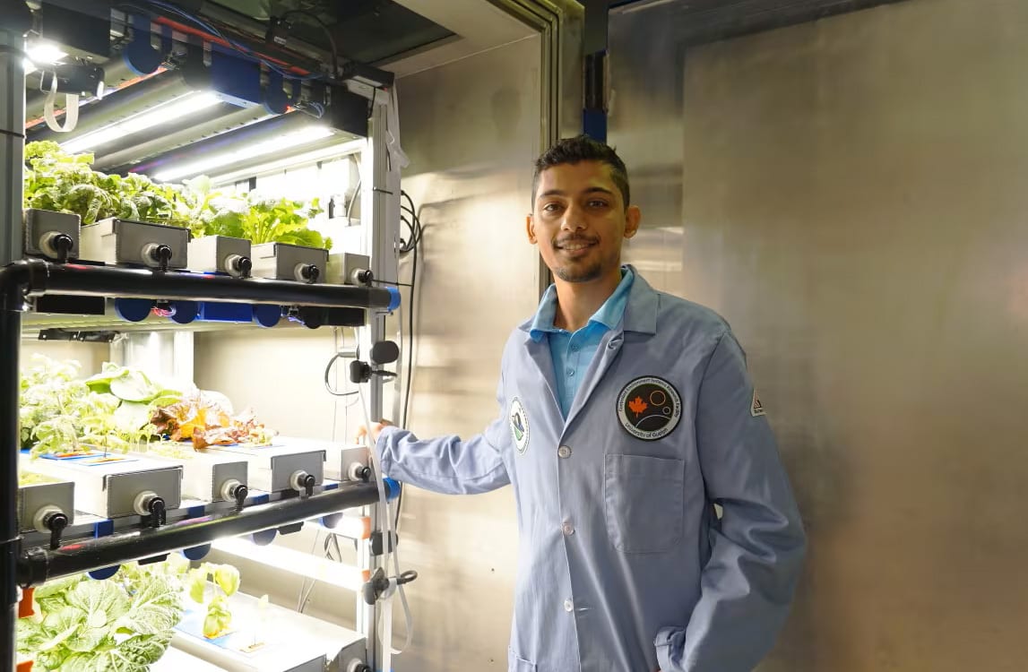 Man in lab coat smiles by a vertical farm with hydroponic plants under LED lights.