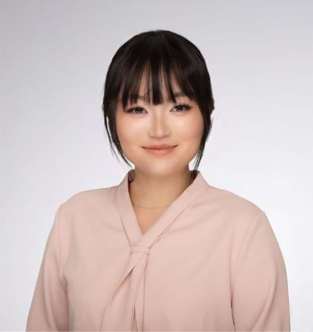 Smiling young Asian woman with dark hair and bangs in a pink blouse, headshot against a gray background.