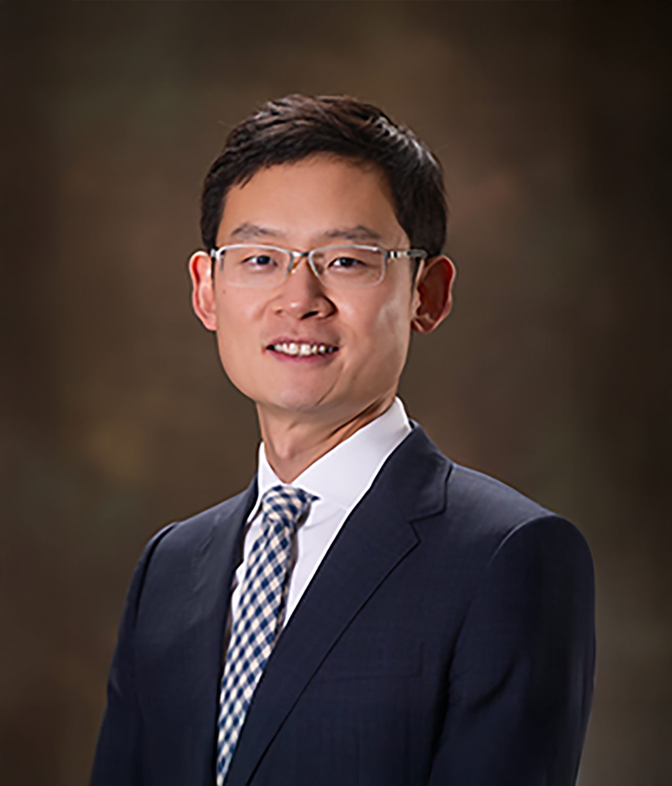 Smiling man with glasses in a dark suit and checkered tie.
