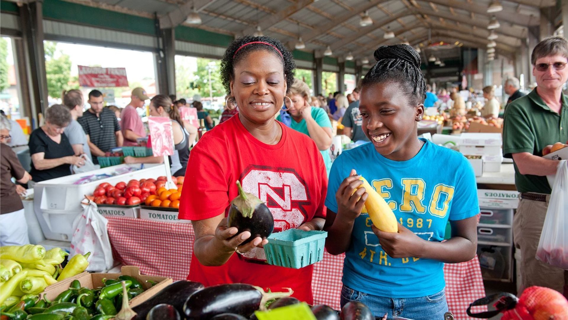 Two smiling Black women at a farmers market, holding an eggplant and yellow squash.