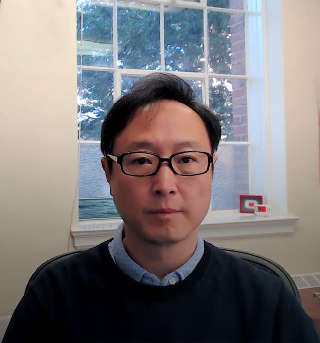A man in glasses and a dark sweater faces the camera, with a window and outdoor view behind him.