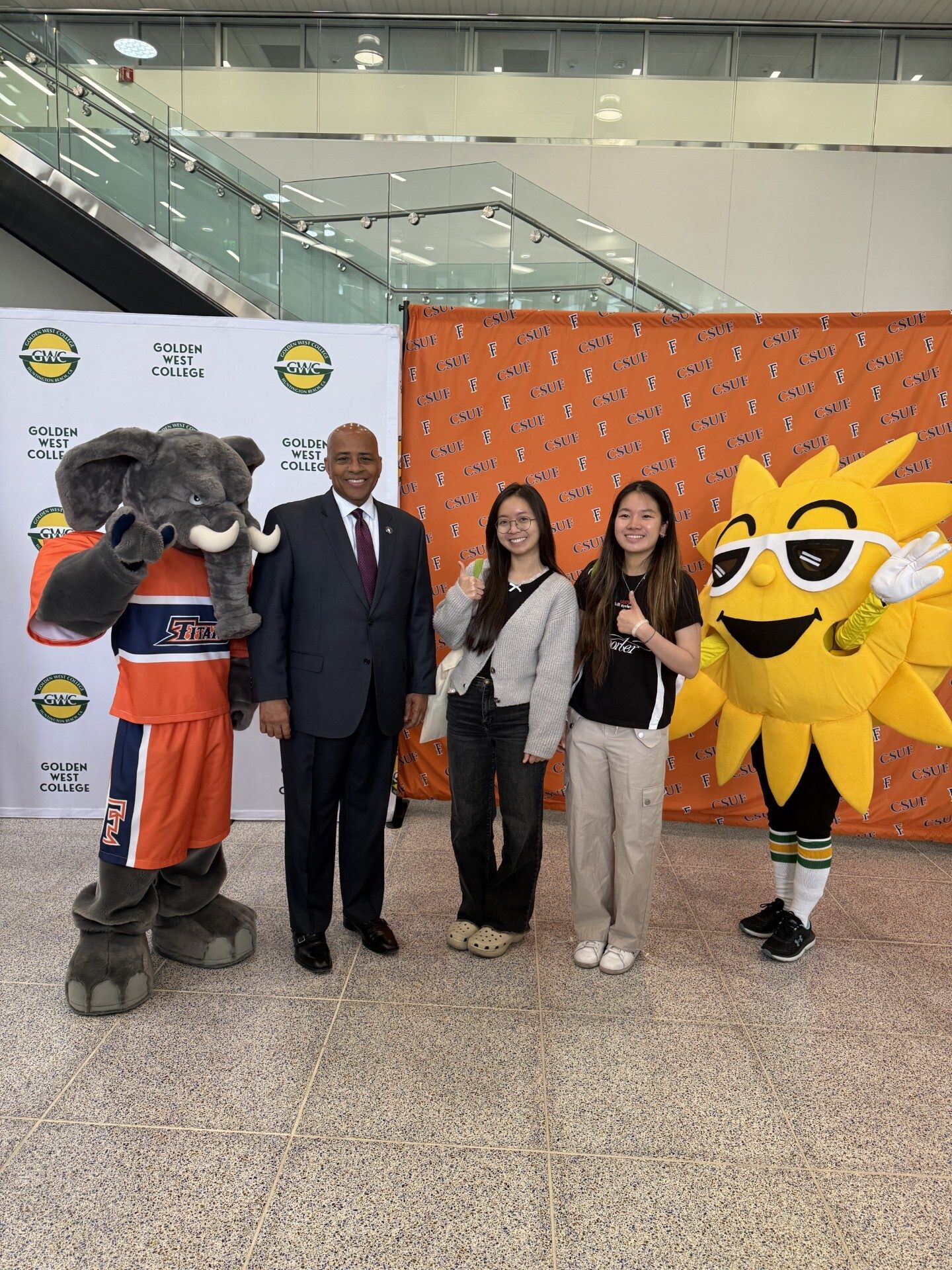 College event photo with two mascots, a man in a suit, and two students giving thumbs up.