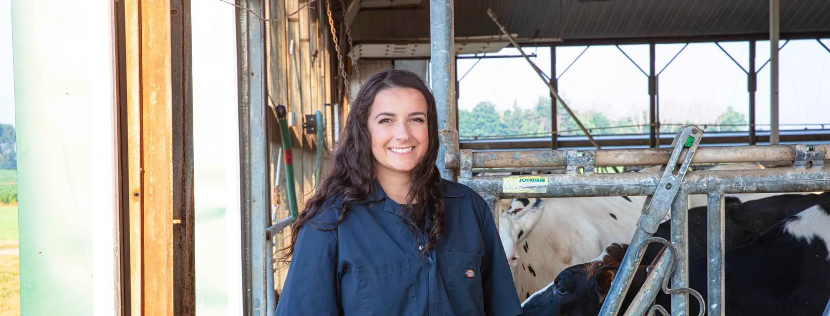 Smiling young woman in a blue work shirt in a barn with cows.