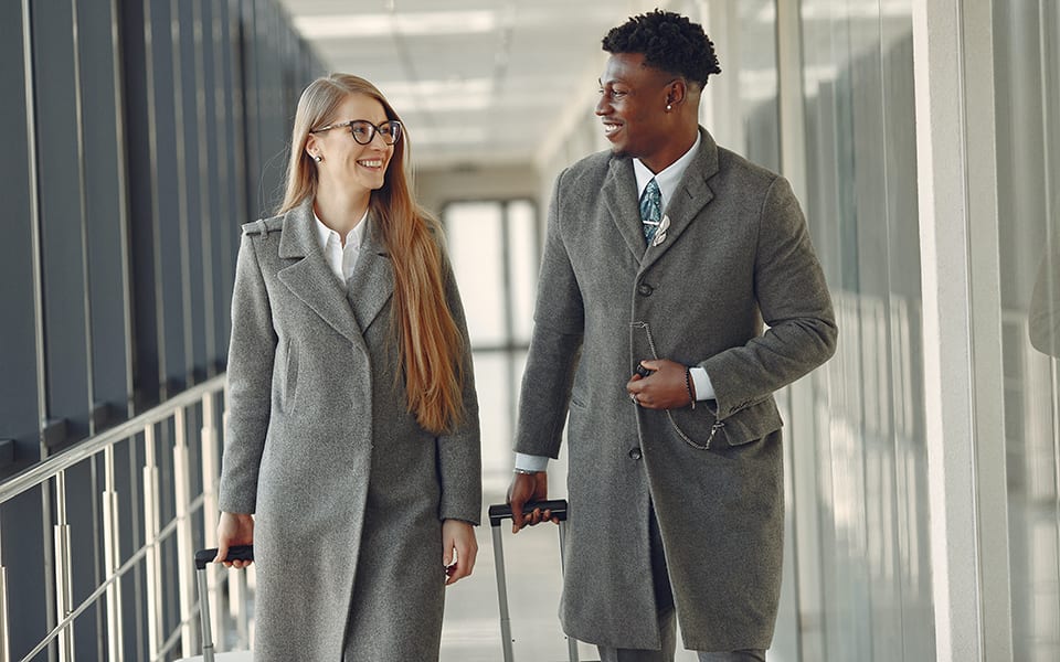 Two business people, a woman and a man, in grey coats smiling and walking with luggage.