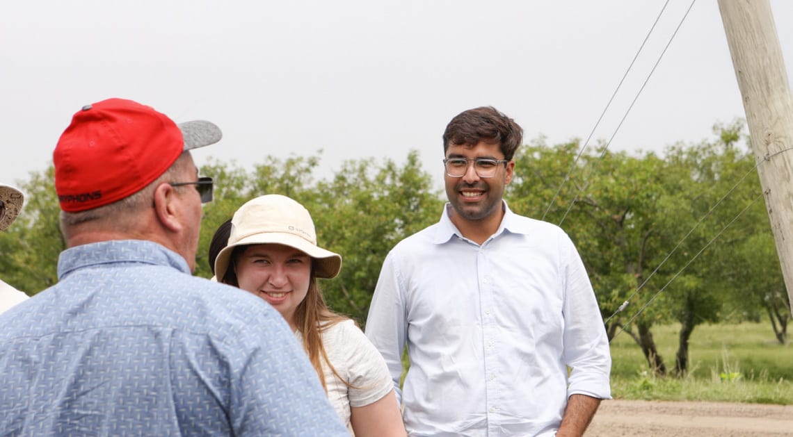 Three people outdoors: a man in a red hat (back to camera), a smiling woman, and a smiling man in glasses.