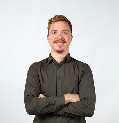 Smiling man with red hair, mustache, dark shirt, arms crossed.