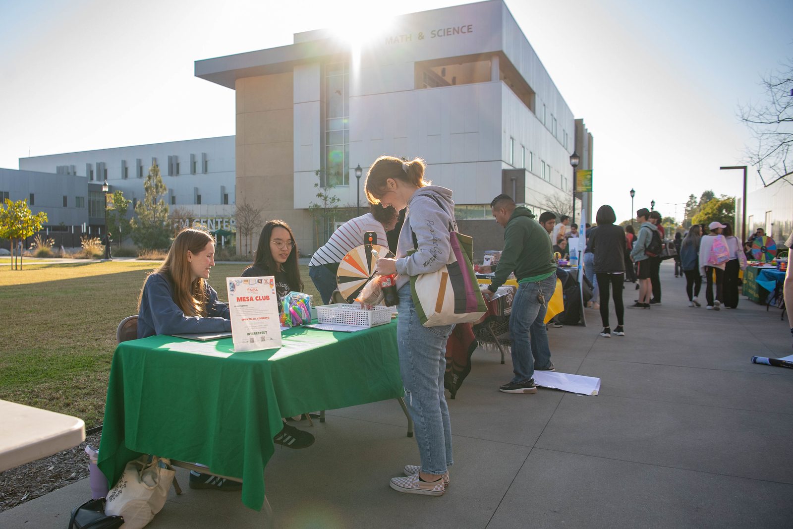 Students at a "MESA Club" booth on a sunny campus, with buildings in the background.