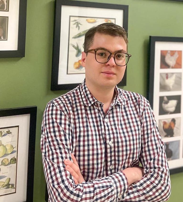 Man with glasses, plaid shirt, arms crossed, by green wall with art.