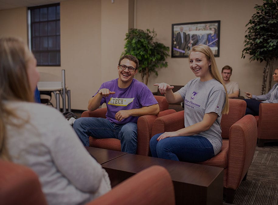 Two university students in a lounge smile and point towards a peer.