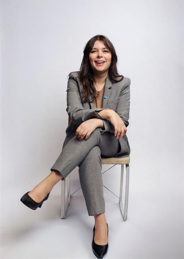 A woman in a grey suit sits on a chair, smiling with her legs crossed and arms folded.