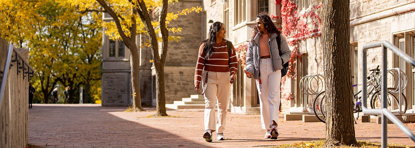 Two women walk on a fall campus path with colorful trees and ivy on buildings.