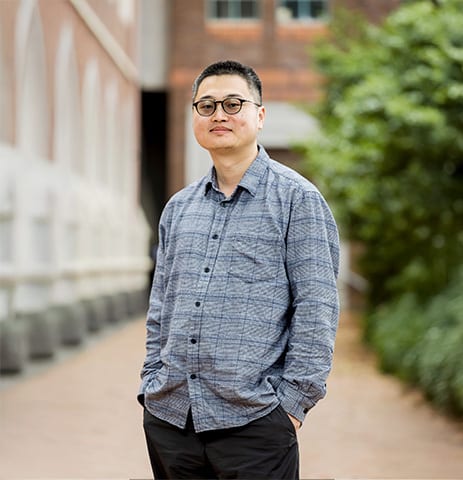 East Asian man with glasses and plaid shirt standing outdoors.