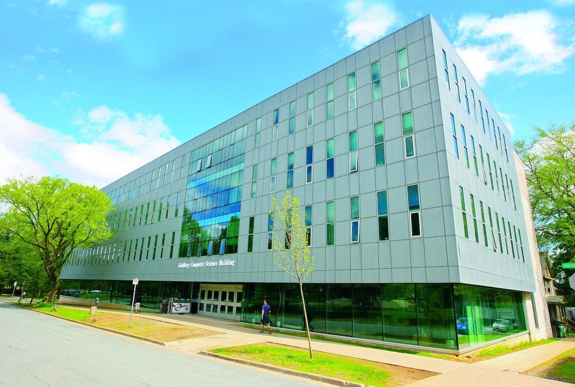 Modern grey Goldberg Computer Science Building with glass windows, trees, street, and blue sky.