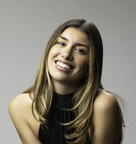A smiling young woman with long brown hair, wearing a black top, looks directly at the viewer.