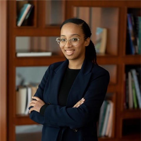 A young woman with glasses and a dark blazer smiles with crossed arms in front of a bookshelf.