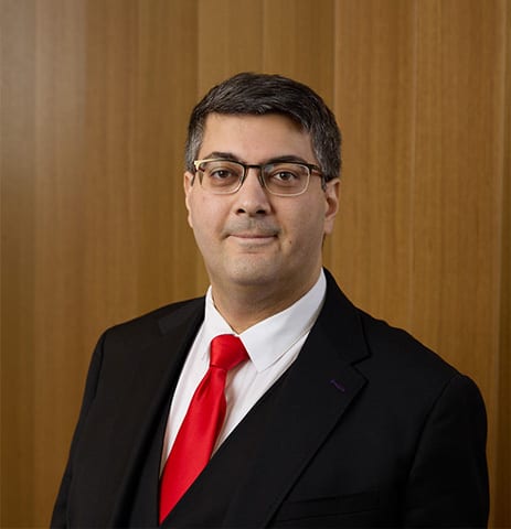 Man in suit, red tie, glasses, smiling, against wood background.