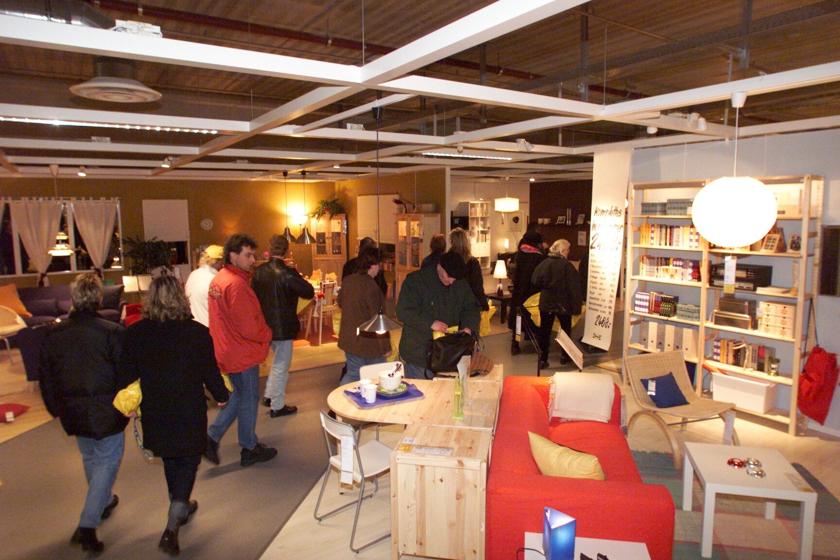 People browse furniture displays in a brightly lit showroom with sofas, shelves, and tables.