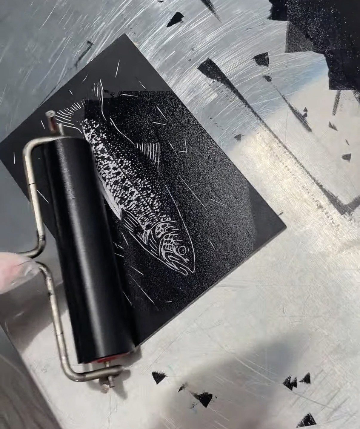A printmaking brayer rolling across a black linocut block featuring a detailed fish design.