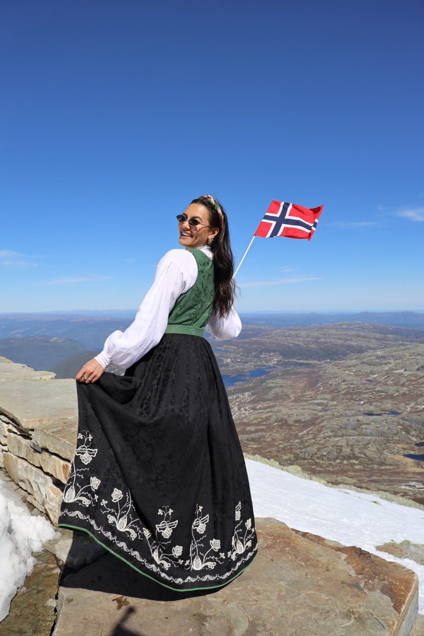 Smiling woman in traditional Norwegian dress holding a flag on a scenic mountain.