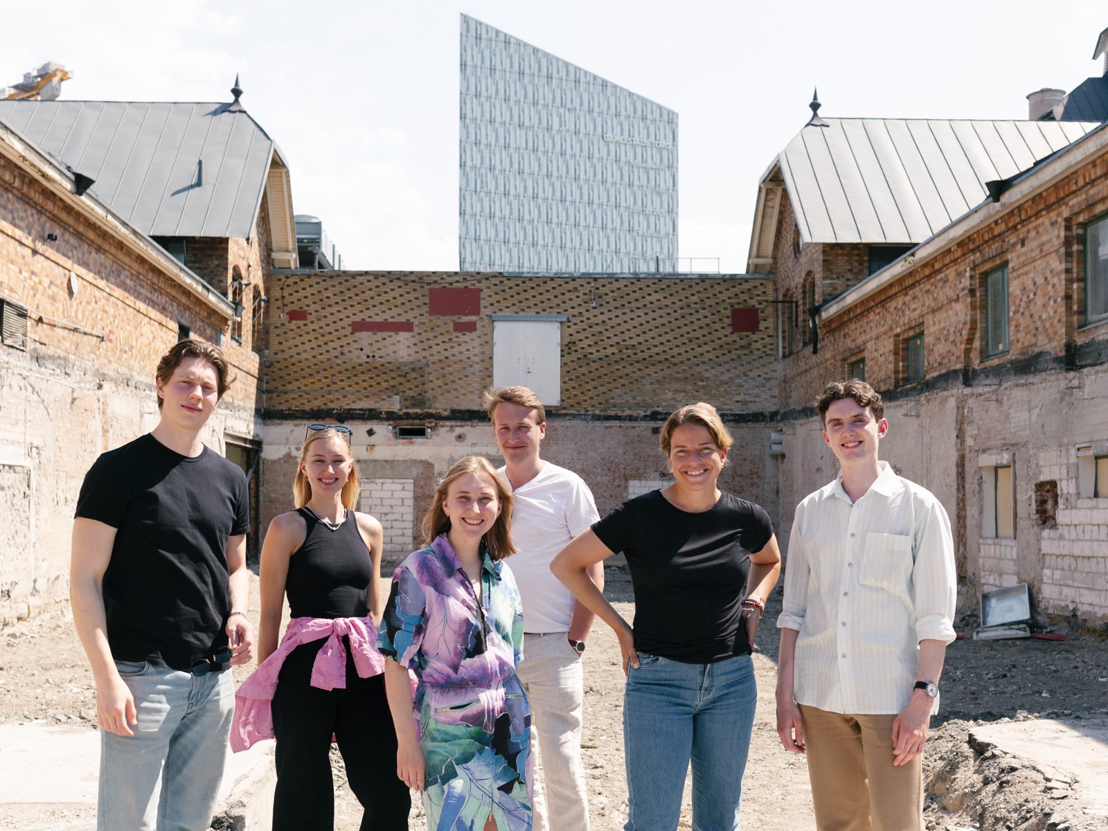 Six smiling young people outdoors with old brick buildings and a modern tower in the background.