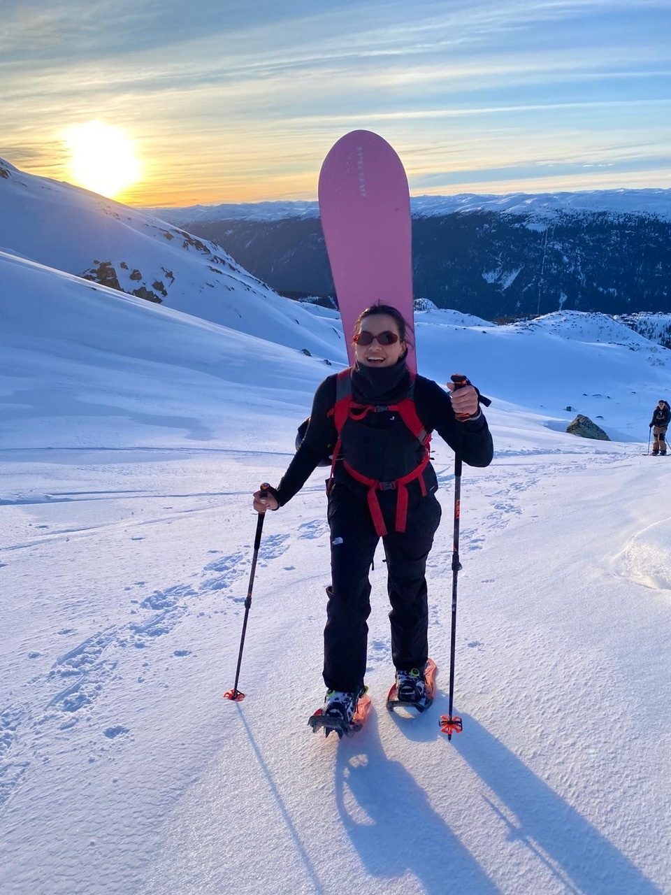 Woman snowshoeing with a pink snowboard on a snowy mountain at sunset.