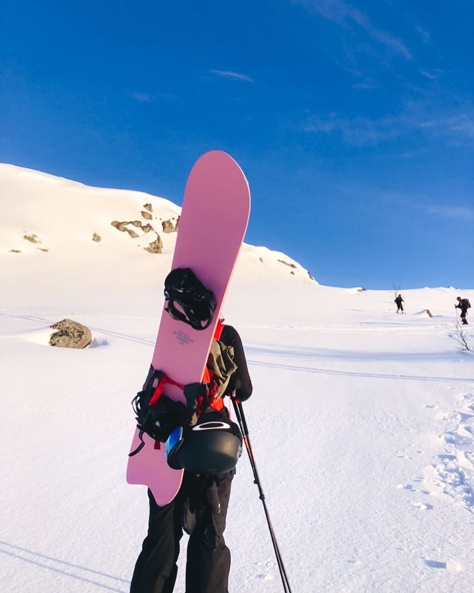 Person carrying a pink snowboard and poles on a snowy mountain under a clear blue sky.