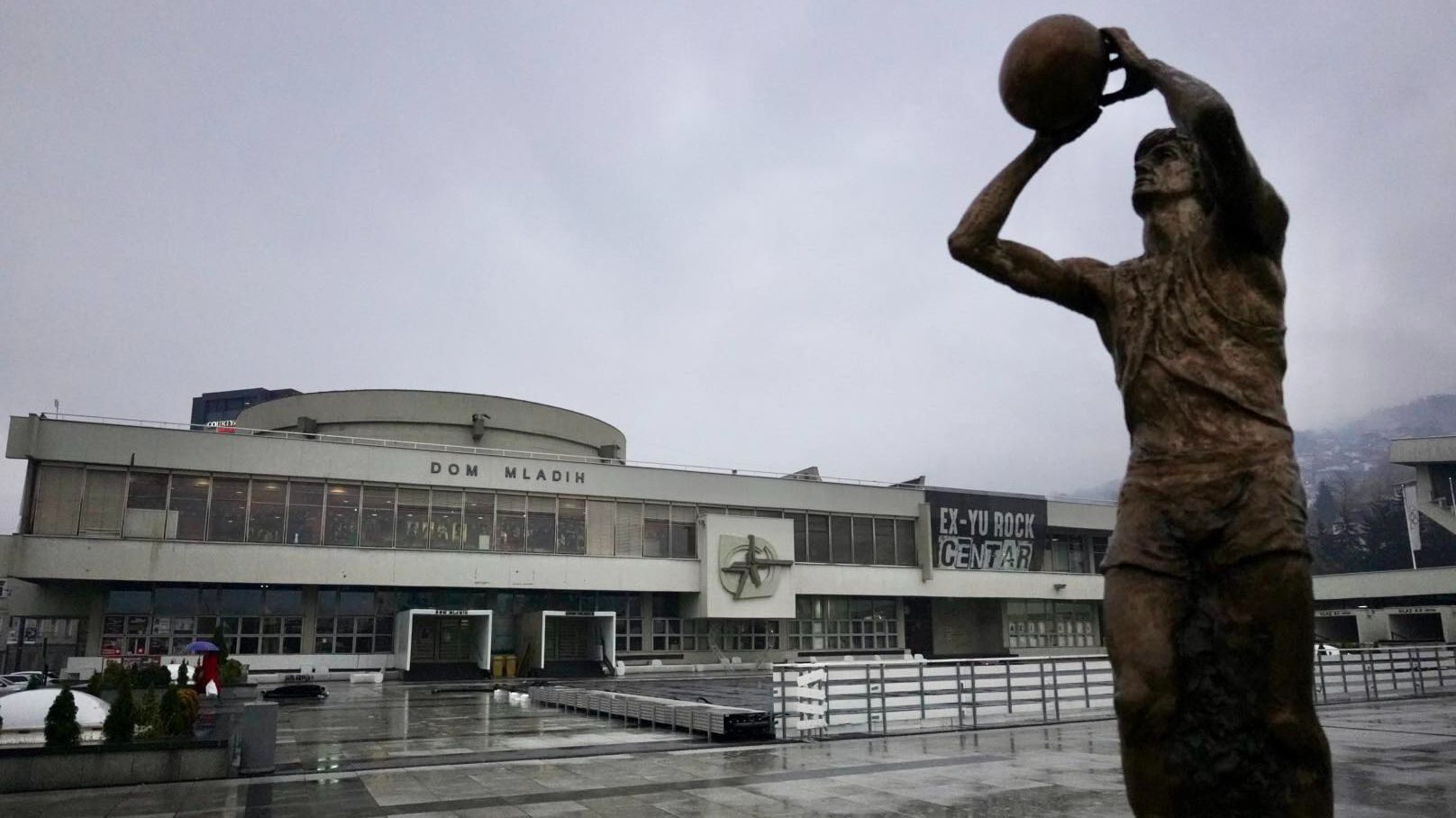 Bronze statue of basketball player and "Dom Mladih" building with "EX-YU ROCK CENTAR" sign under overcast sky.