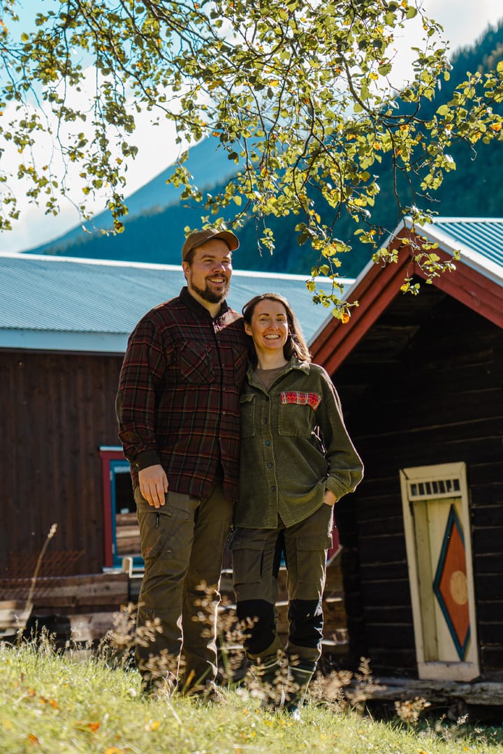 Happy couple outdoors, standing near rustic buildings with mountains in background, leaves above.