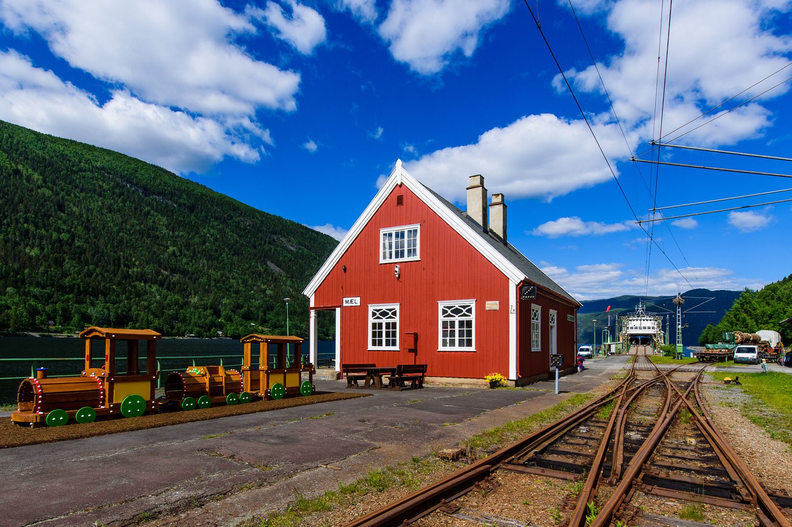Red train station by a lake with tracks, a wooden play train, and green mountains under a blue sky.