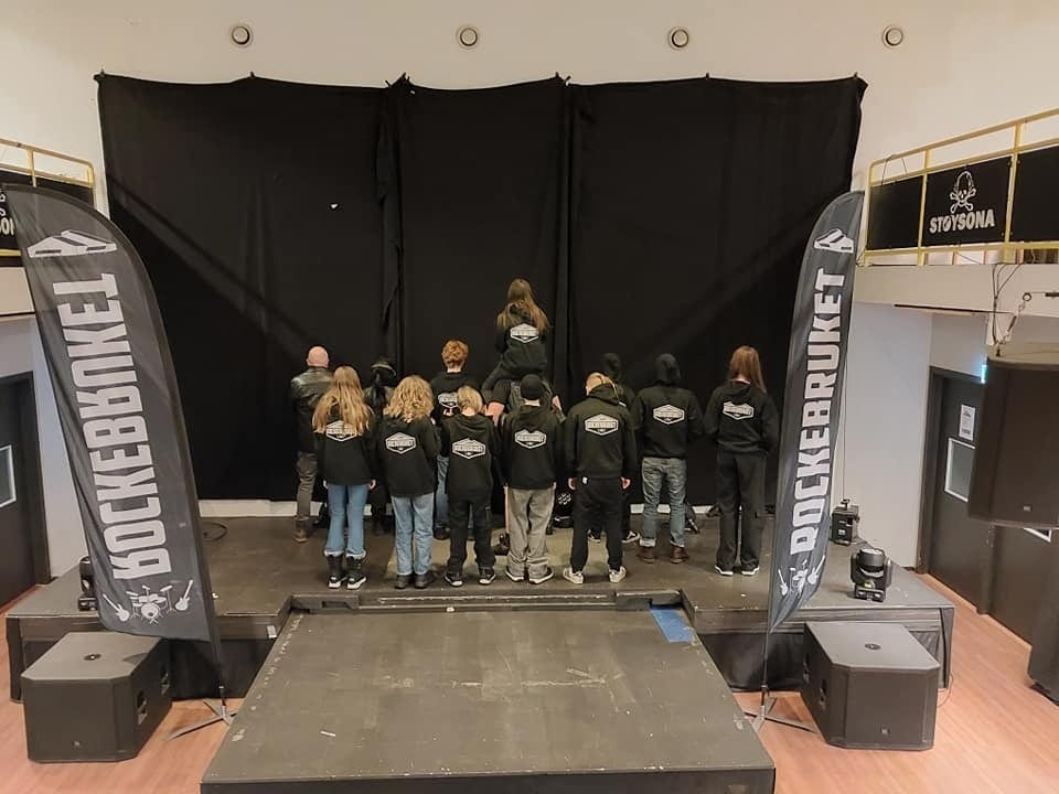 Group facing a black backdrop on a stage, wearing black hoodies, with two "ROCKEBRUKET" banners.