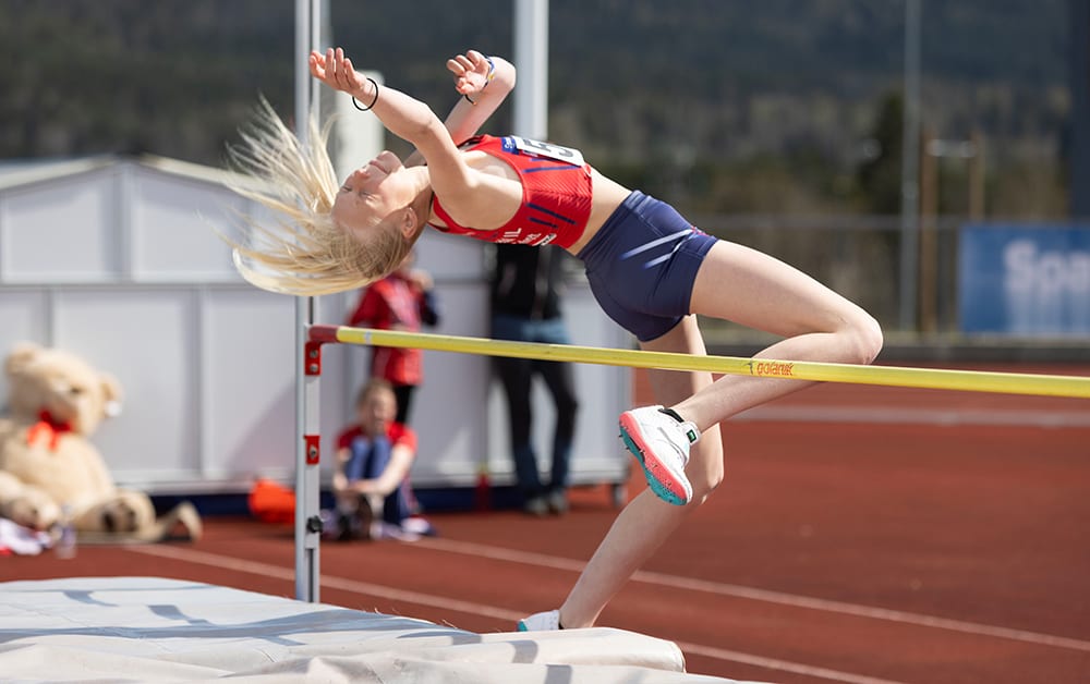 Blonde female high jumper clears the bar backwards, mid-air, with flowing hair.